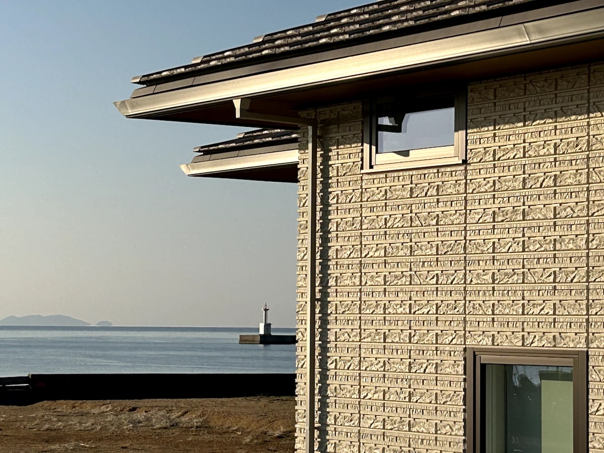 The exterior of the newly constructed rental villa is featured, showcasing textured wall panels and a sloping roof. In the background, the tranquil waters of Osaka Bay are visible, along with a lighthouse standing at a distance, enhancing the coastal scenery.