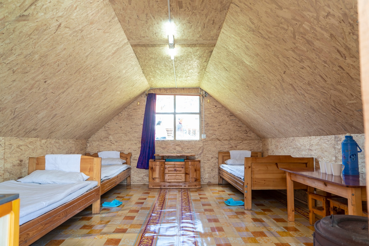 An attic-style room features two wooden beds with white linens positioned on either side. A wooden storage cabinet is centrally placed. A window brings in natural light, complemented by a dark curtain. A wooden table holds blue towels and a drink dispenser.