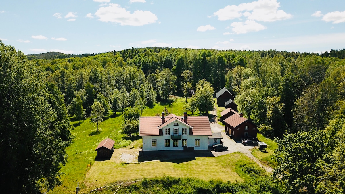 An aerial view of a spacious property set amidst lush green landscapes. The main building features a distinctive roof and is surrounded by trees, with additional smaller structures and open grassy areas visible nearby.