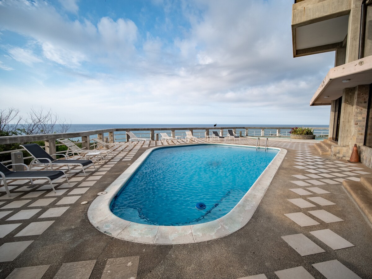 An outdoor pool is situated on a tiled patio, offering views of the ocean in the background. Several lounge chairs are arranged around the pool area, while the sky is partially cloudy, enhancing the serene ambiance.