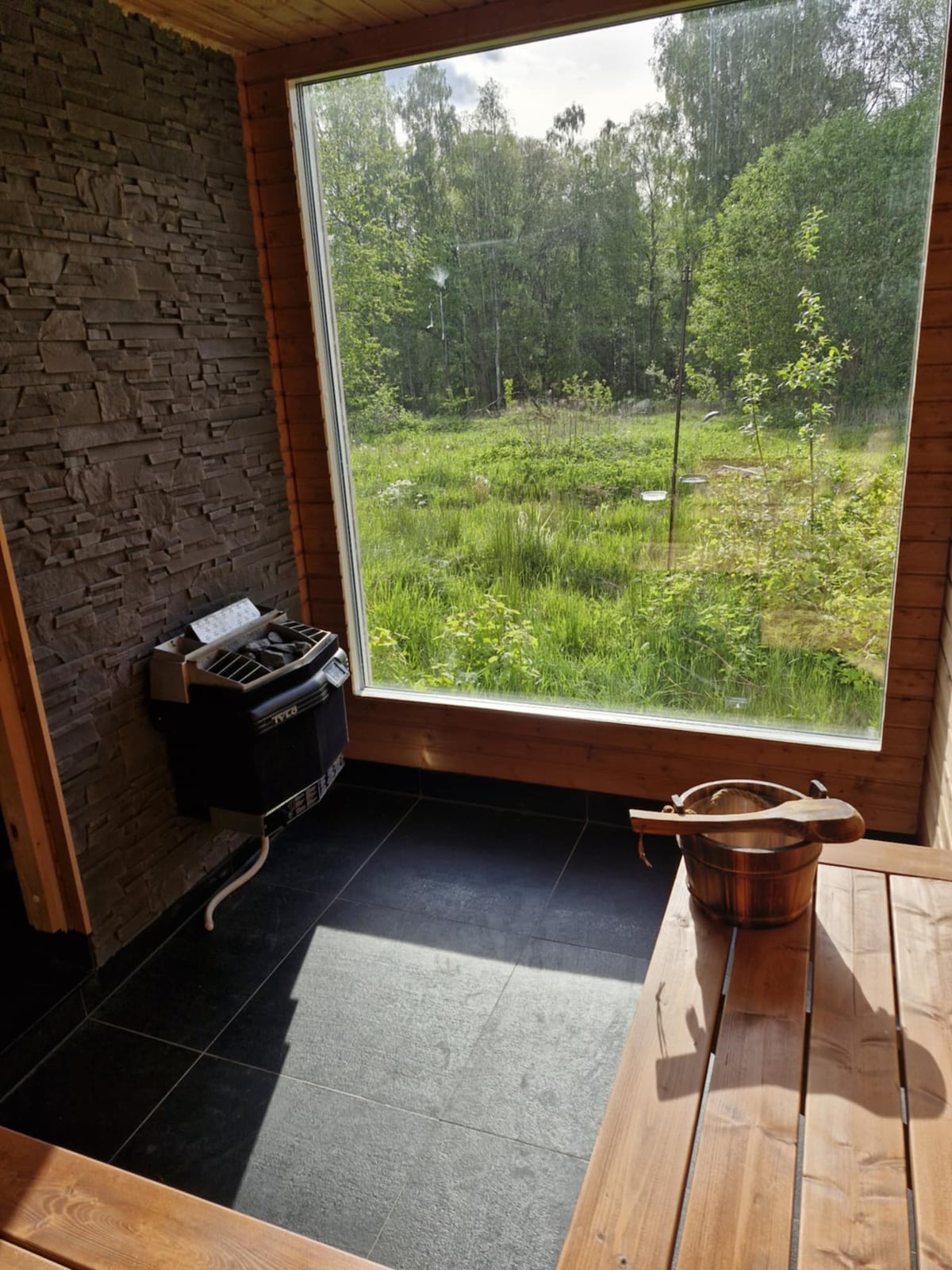 A sauna space features a large window overlooking a lush green landscape. A wooden bench is positioned alongside a traditional sauna stove, and a bucket is placed on the bench, contributing to the authentic sauna experience.