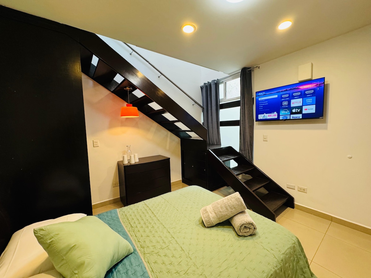 A cozy sleeping area features a spacious bed with light green bedding. A sleek dresser is positioned near the foot of the bed. A wall-mounted Smart TV is visible, with a staircase leading to an upper level. Soft lighting enhances the calming atmosphere.