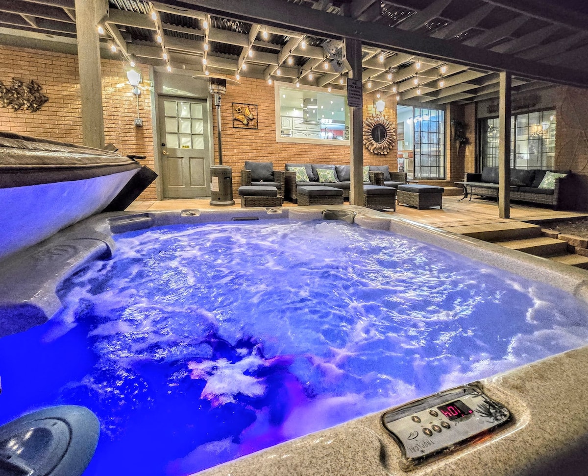 A relaxing hot tub is showcased, illuminated by soft blue lights, inviting relaxation. The spacious outdoor patio features comfortable seating, perfectly blending indoor and outdoor living. Walls lined with brick provide a cozy ambiance, while soft lighting highlights the inviting gathering space.