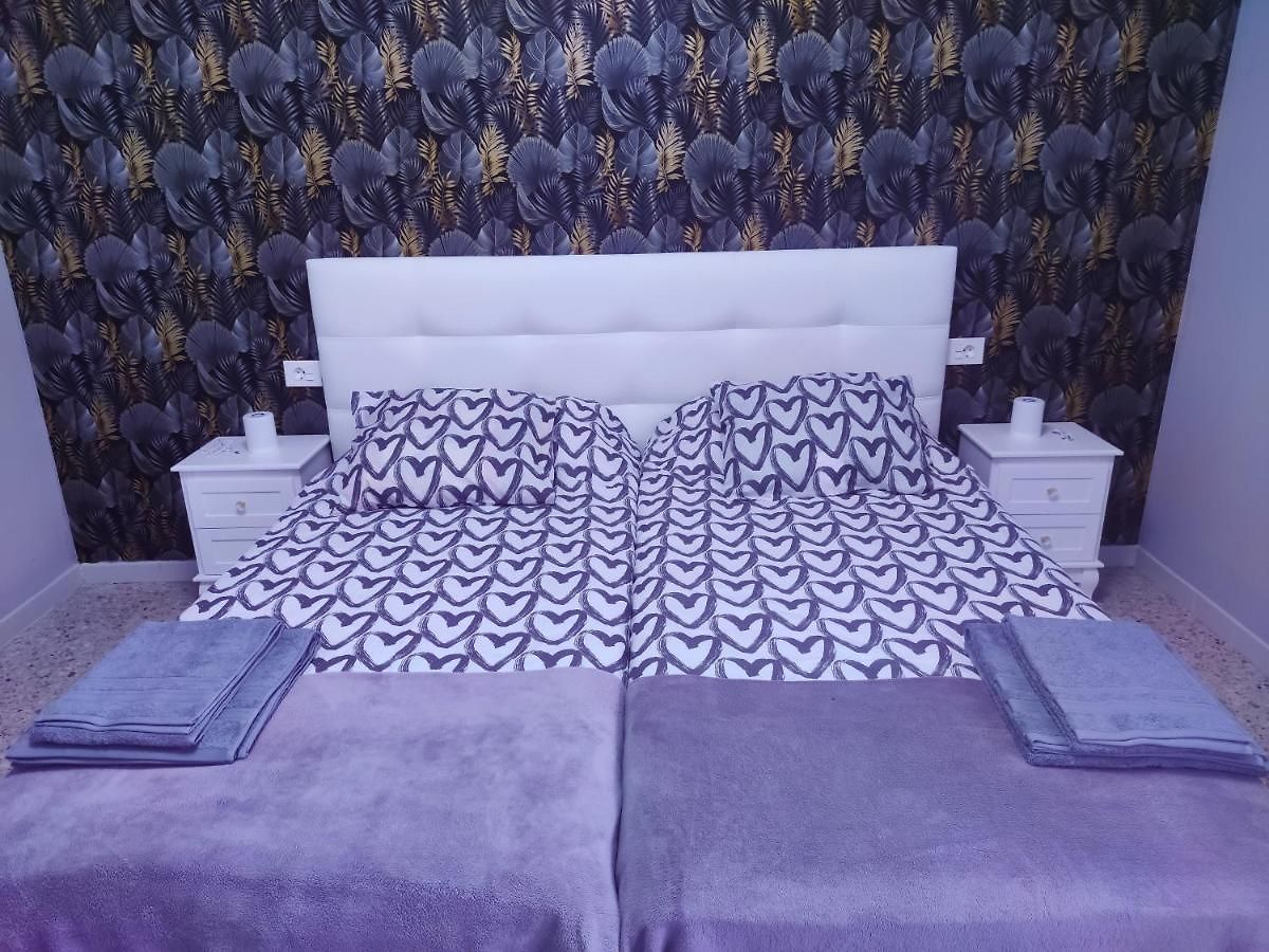 A spacious sleeping area features a white bed frame with a patterned bedspread in heart motifs. Two bedside tables are positioned on either side, each with a small lamp. Soft towels in a muted shade are neatly placed on the bed, complementing the stylish wallpaper with leaf designs.