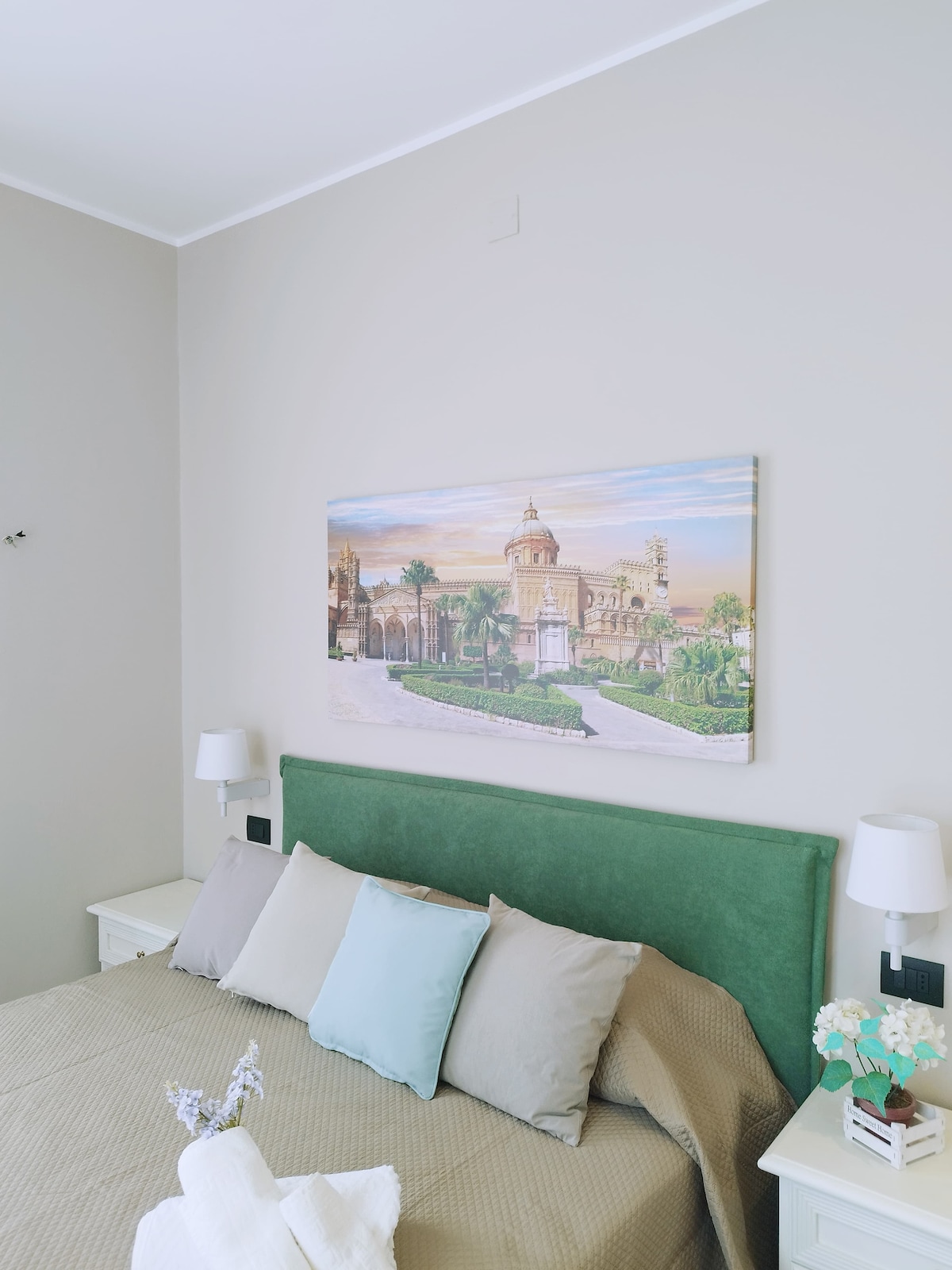 A serene bedroom features a plush green headboard with a soft, neutral-colored bedspread. Decorative pillows in various shades provide a subtle accent. A large landscape painting depicting an ornate structure is mounted above the headboard, while bedside lamps offer gentle illumination.