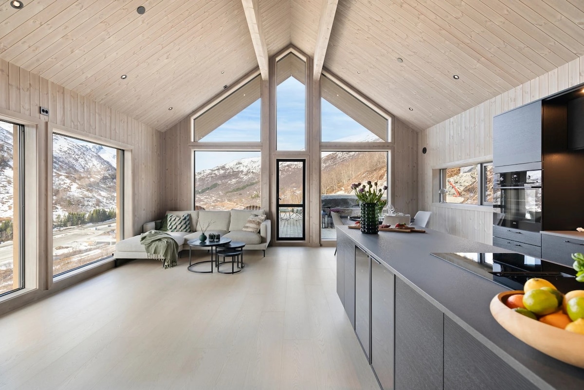 A modern living area is showcased with high ceilings and large windows offering expansive views of the surrounding mountains. A light-colored sofa is positioned beside a stylish coffee table, while a sleek kitchen island features a bowl of fresh fruit. Natural light fills the space.