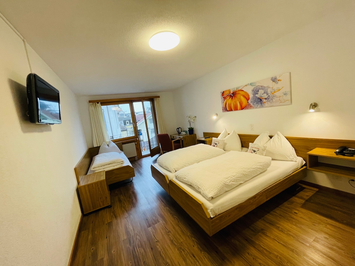 A well-lit room features a spacious bed with white bedding, accompanied by a single bed adjacent to it. A flat-screen TV is mounted on the wall. Light wood furniture and a colorful wall art piece add interest. A balcony door allows natural light inside.