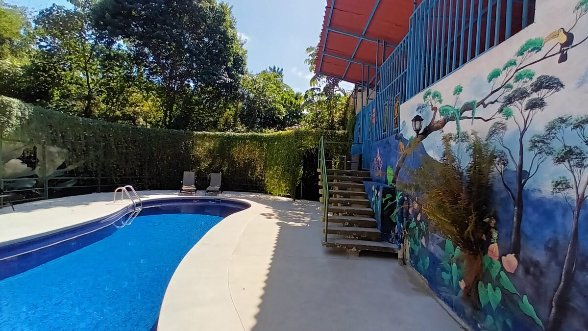 A vibrant mural decorates the exterior of the building adjacent to a refreshing swimming pool. The pool area is surrounded by lush greenery, with comfortable lounge chairs positioned nearby. Stairs lead up to the entrance of the building, enhancing the overall outdoor aesthetic.