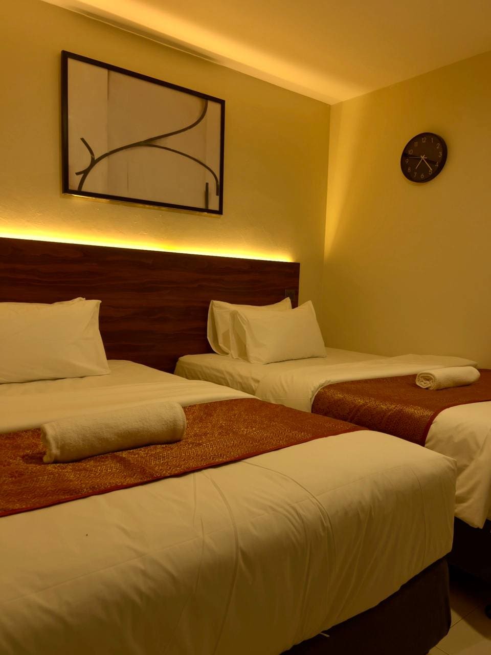 A well-lit bedroom features two spacious beds, each adorned with neatly arranged linens. Soft, warm tones are present in the decor, complemented by a minimalistic wall art piece and a wall clock. A rolled towel rests on the bed for convenience.