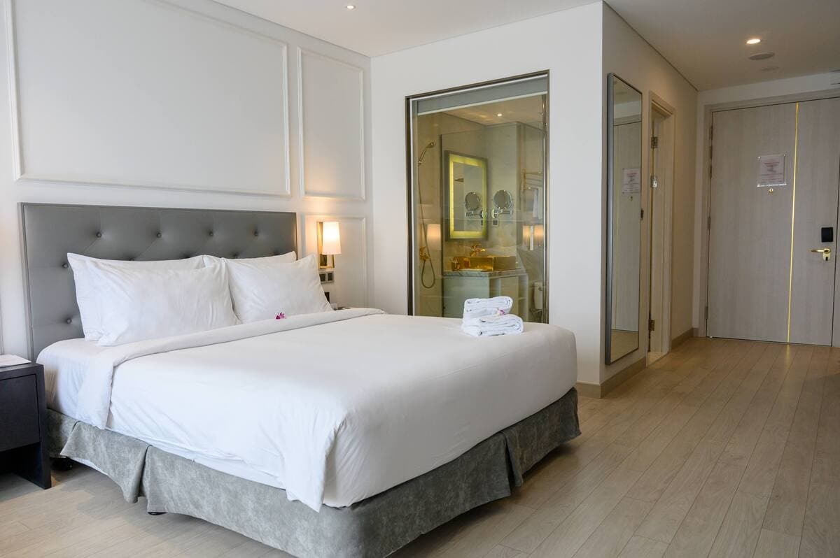A modern hotel room features a spacious bed with fresh bedding, neatly arranged towels, and soft lighting. A private bathroom is visible through glass doors, showcasing a clean design. The light hardwood flooring complements the contemporary decor, providing an open and inviting atmosphere.