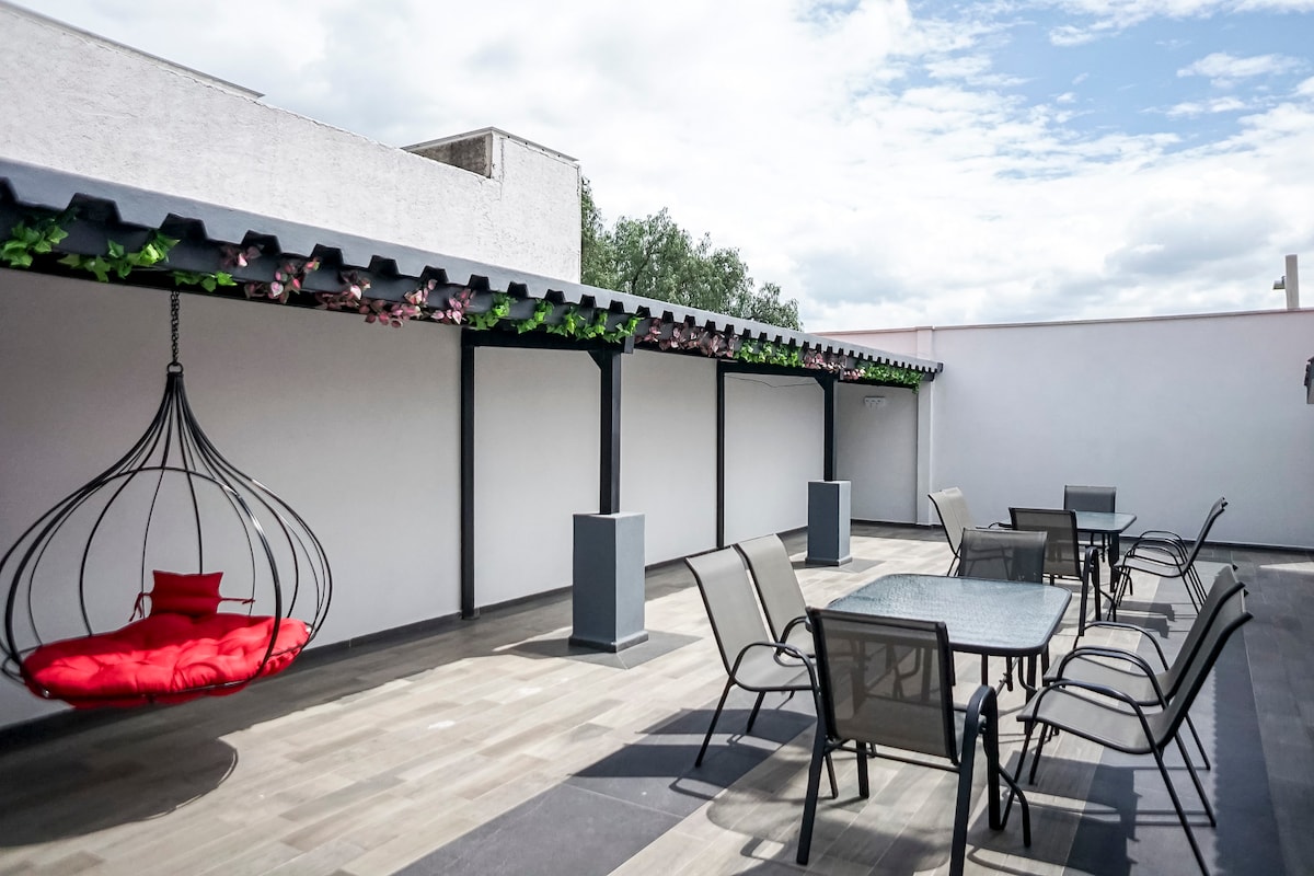 An outdoor terrace features a hanging chair with a red cushion, surrounded by several tables and chairs. The space is shaded by a roof supported by posts, creating a comfortable area for relaxation or socializing. Greenery is present along the edge, adding a natural touch.