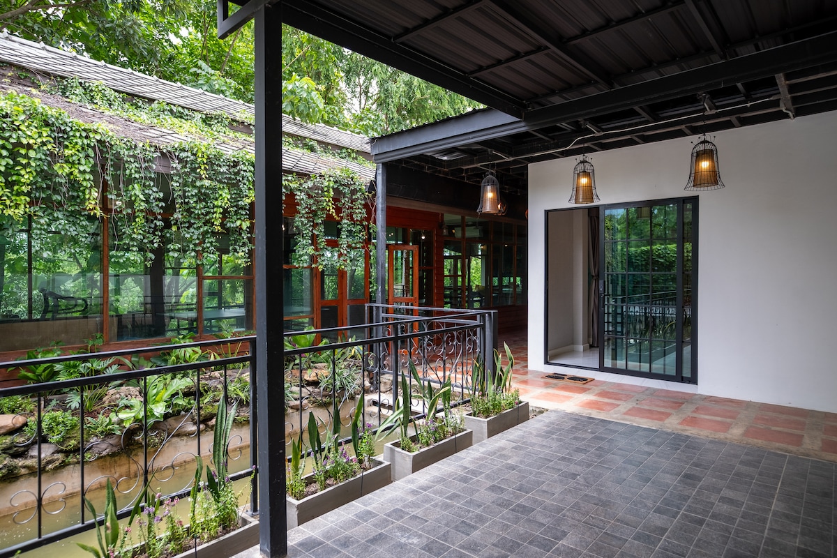 An outdoor patio area is shown, featuring a stone flooring and a metal railing adorned with plants. Natural light is present, illuminating a nearby room with large glass windows. Lush greenery lines the edges, enhancing the tranquil setting.