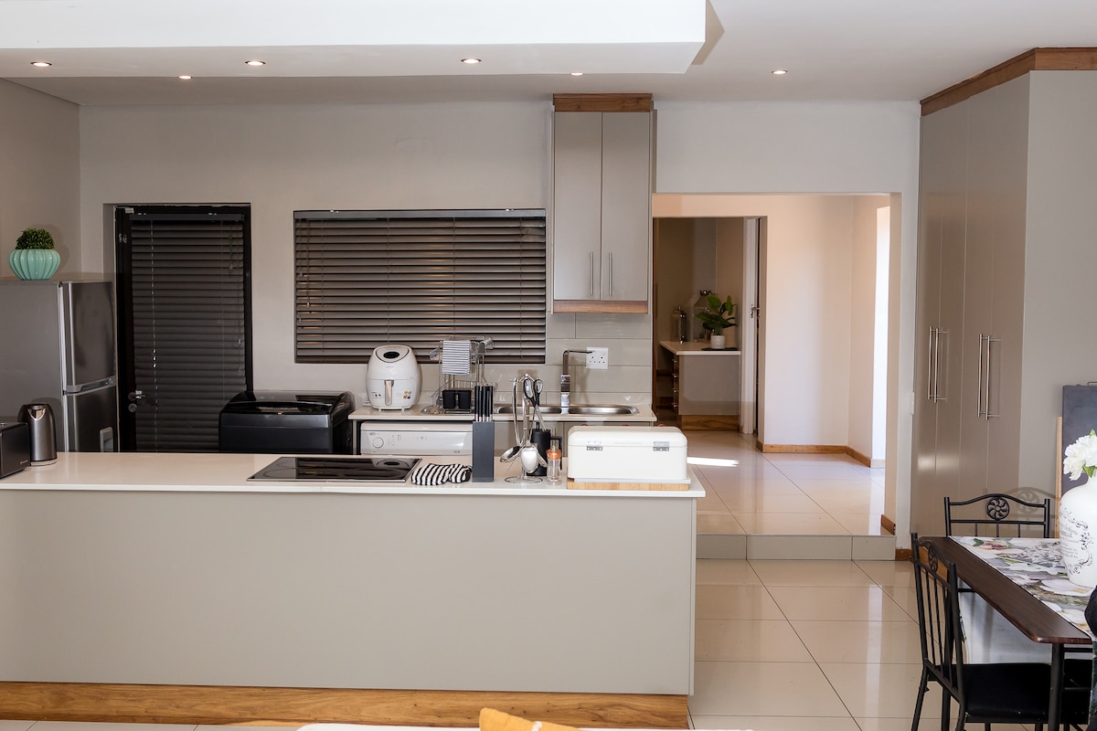 A modern kitchen features sleek cabinetry and a well-equipped countertop. Appliances including a refrigerator and microwave are readily visible. The open layout leads to a dining area, enhancing the spacious feel of the interior. Natural light illuminates the room through large windows.