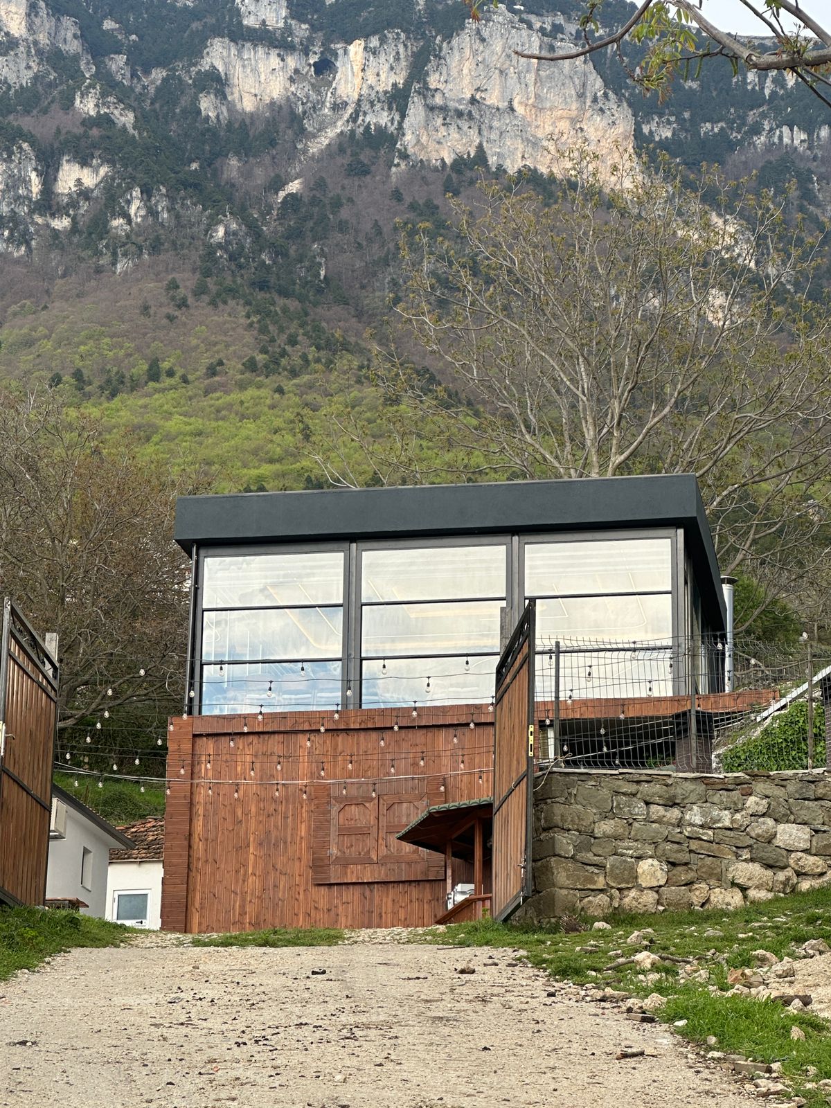 A modern, multi-level building with large glass windows offers views of surrounding greenery and mountain landscapes. The structure features a wooden entrance and is set on a gravel path leading through an outdoor area with stone walls and natural elements.