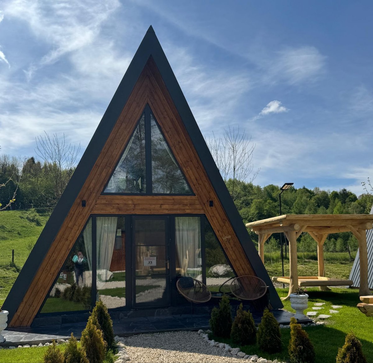 A triangular cabin structure features large glass windows framed by wooden accents. A sandy pathway leads to the entrance, bordered by small shrubs. A sheltered outdoor seating area is visible to the right, creating an inviting space to enjoy the natural surroundings.