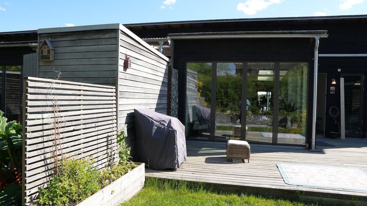 A modern outdoor area is shown, featuring a wooden deck with a seating arrangement. A covered outdoor grill is positioned near a low planter filled with greenery. Large glass doors lead into the interior space, surrounded by a serene landscape.