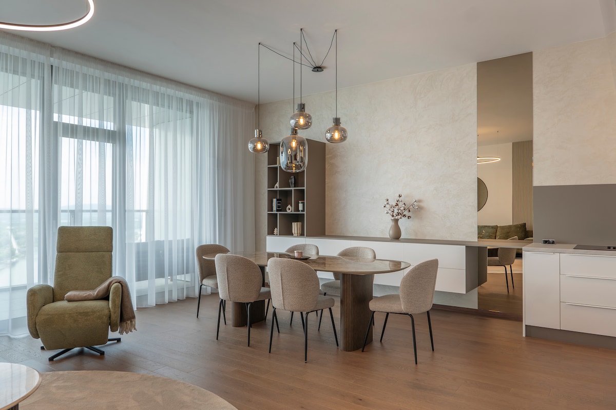 A light-filled living area features a round dining table with soft, upholstered chairs. A green armchair is positioned nearby, complemented by large windows draped in sheer curtains. Modern pendant lighting enhances the clean lines of the space, promoting a welcoming atmosphere.