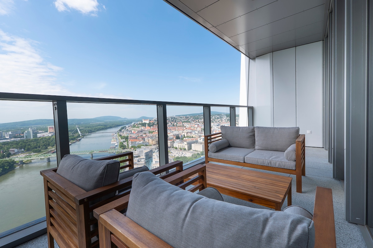 A balcony seating area is furnished with wooden furniture, including a table and multiple sofas, providing ample space for relaxation. The panoramic view displays the river and cityscape, with clear skies enhancing the outdoor experience.