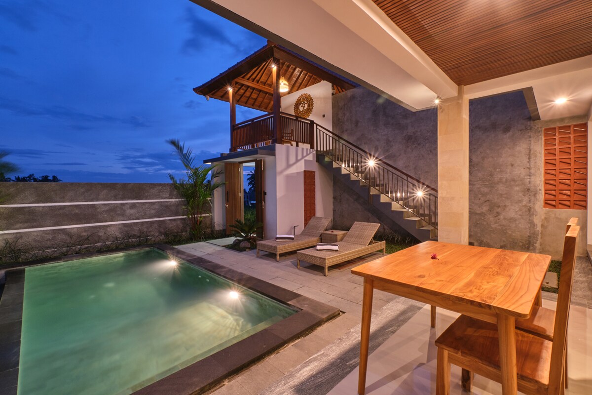 An inviting outdoor space features a rectangular swimming pool surrounded by sleek stone tiles. Two lounge chairs are positioned nearby, alongside a wooden dining table with upright chairs. A staircase leads to a second-level area, accentuated by a traditional Balinese roof and soft evening lighting.