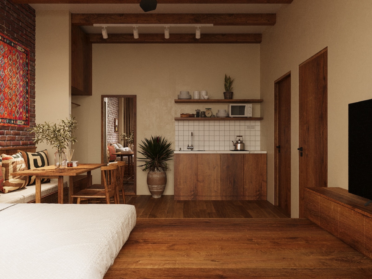 A warm and inviting studio apartment features wooden beams and a light color scheme. A comfortable large bed is positioned near a compact kitchen equipped with essential appliances. A dining table with chairs is next to a sitting area adorned with decorative cushions.