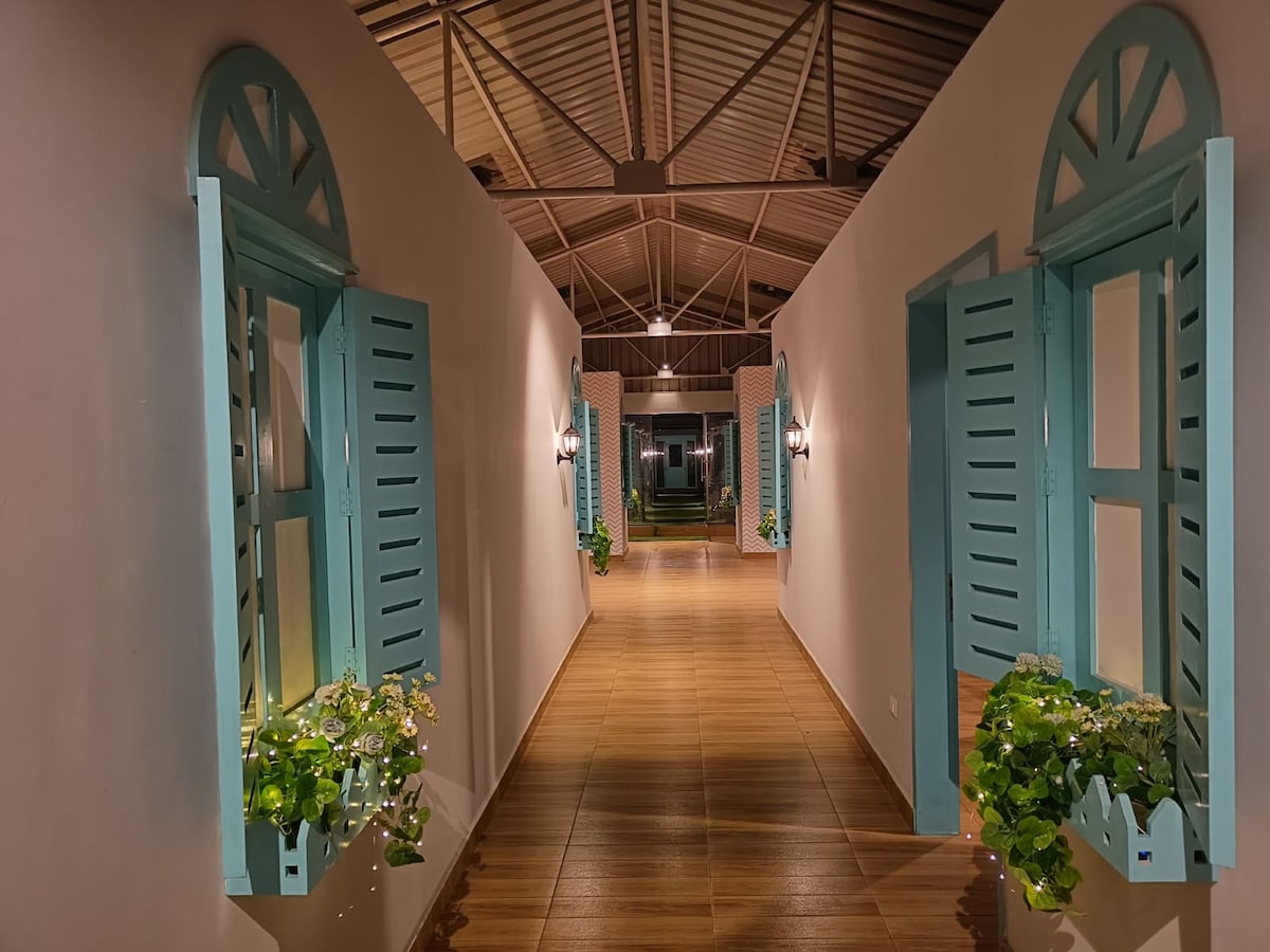 An elongated hallway is lined with open turquoise shutters, each accompanied by potted greenery. Warm lighting emanates from wall sconces, and wooden flooring runs throughout, leading to a well-lit area at the end. The high ceiling features a metal framework, enhancing the spacious feel.