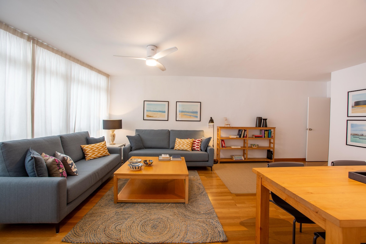 This living area features two gray sofas with colorful cushions, oriented towards a central wooden coffee table. A light-colored rug sits beneath the seating arrangement. Natural light is provided by large windows draped with sheer curtains, and a ceiling fan contributes to the airy ambiance.