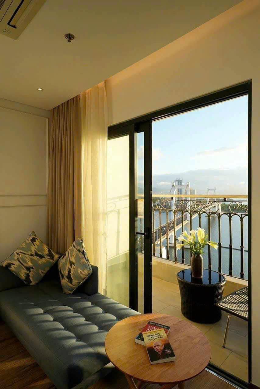 A comfortable seating area features a sleek, dark sofa adorned with patterned cushions. A small wooden table holds a stack of magazines. Floor-to-ceiling glass doors open to a balcony, offering views of the surrounding landscape.