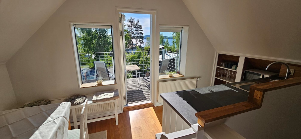 Natural light fills the open-plan living space, highlighted by large windows offering views of the lake. A comfortable seating area is complemented by wooden flooring, while a small dining table is visible on the balcony, inviting outdoor meals.