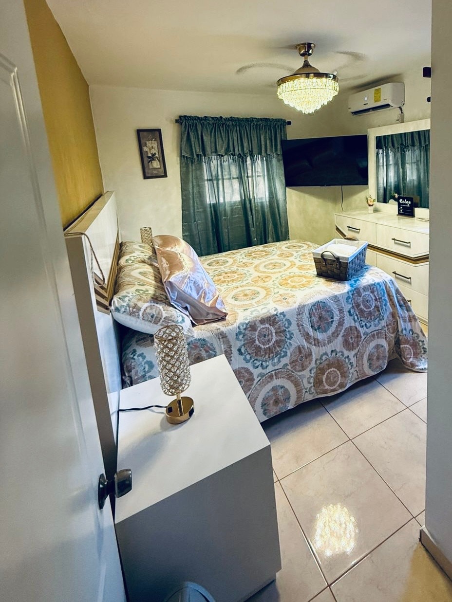 A cozy bedroom is featured, with a spacious bed adorned with a patterned blanket. Soft curtains frame the window, allowing natural light to enhance the space. A television is mounted on the wall opposite the bed, and a small bedside table holds a decorative lamp.