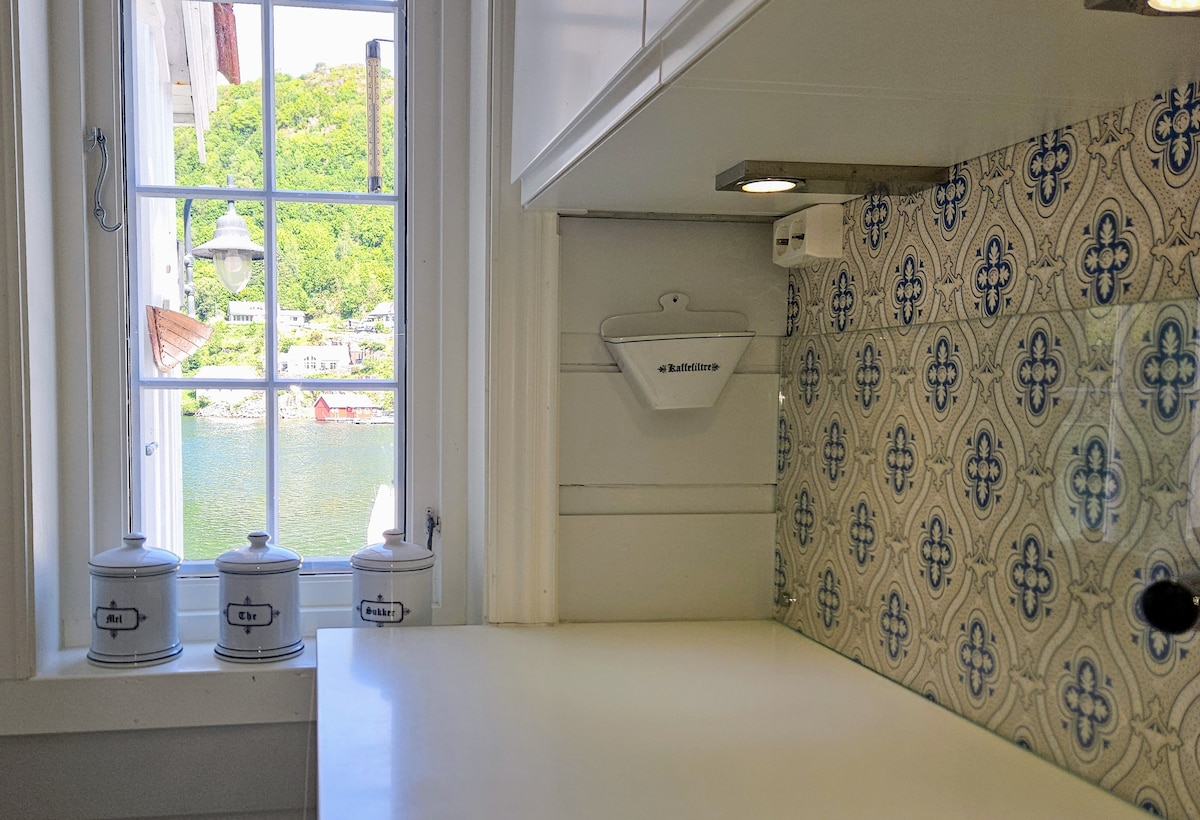 A bright kitchen area features a window offering a view of the water and surrounding greenery. Decorative tiles with blue patterns line the wall. Three canisters are neatly arranged on the counter, complementing the clean, minimalist design.
