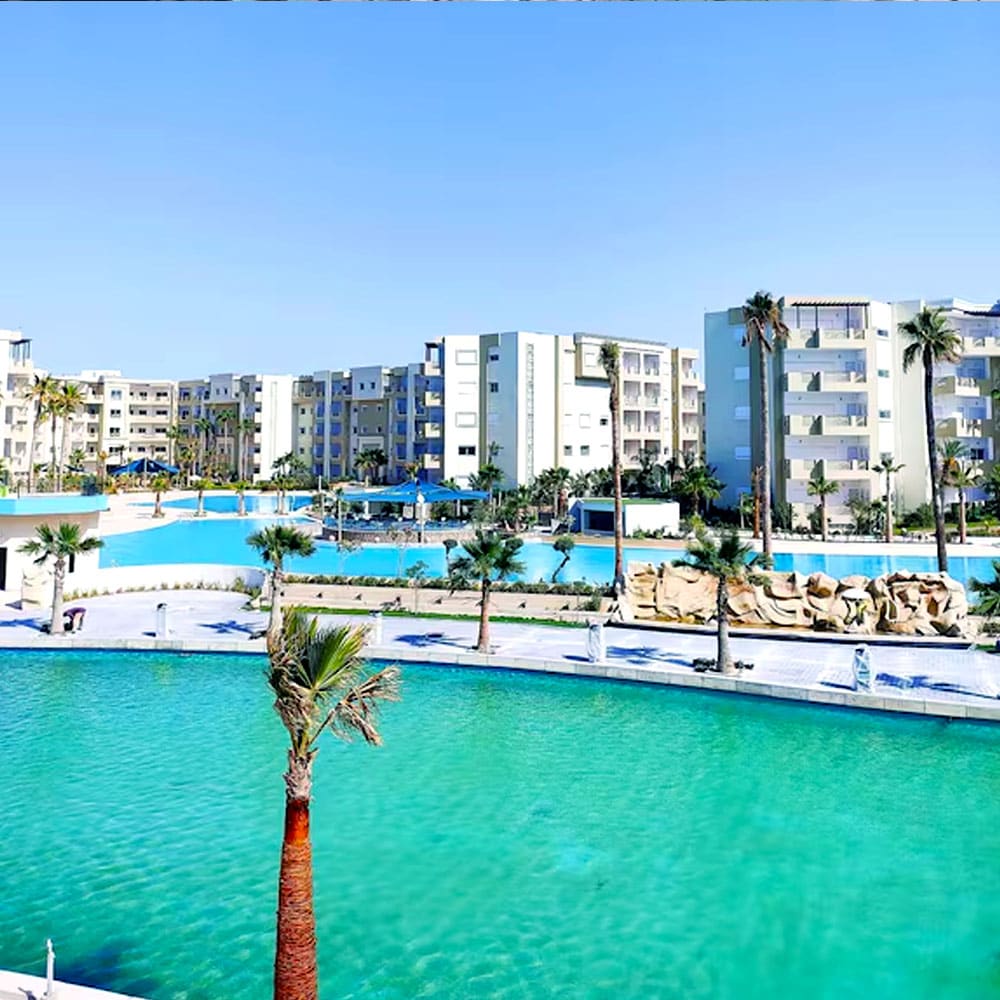 A broad view of the expansive swimming pools is depicted, surrounded by lush palm trees. The modern buildings of the residence are visible in the background, alongside inviting blue waters that create a refreshing atmosphere.