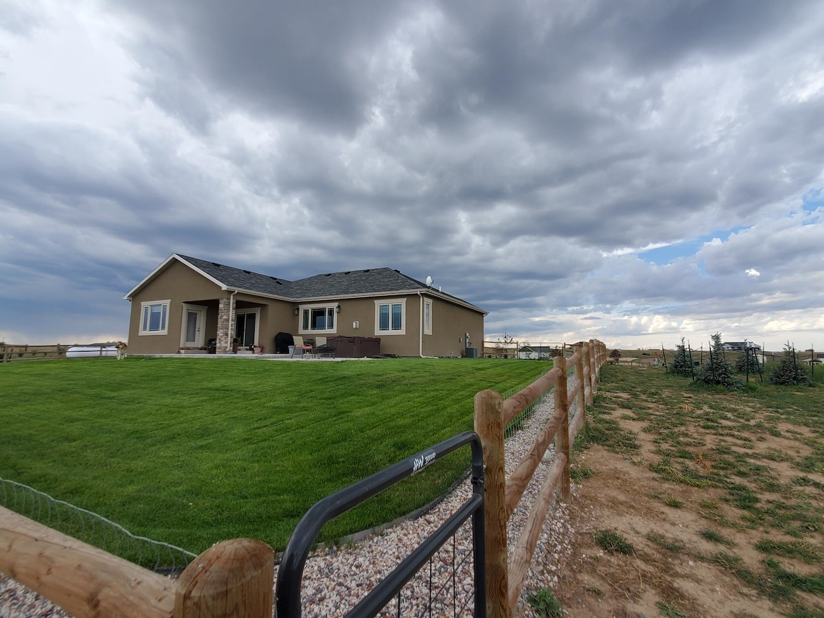 A single-story home is set amidst a spacious green lawn, surrounded by a wooden fence. The structure features several large windows and a covered patio area. A cloudy sky looms above, creating a serene backdrop to the peaceful country retreat.