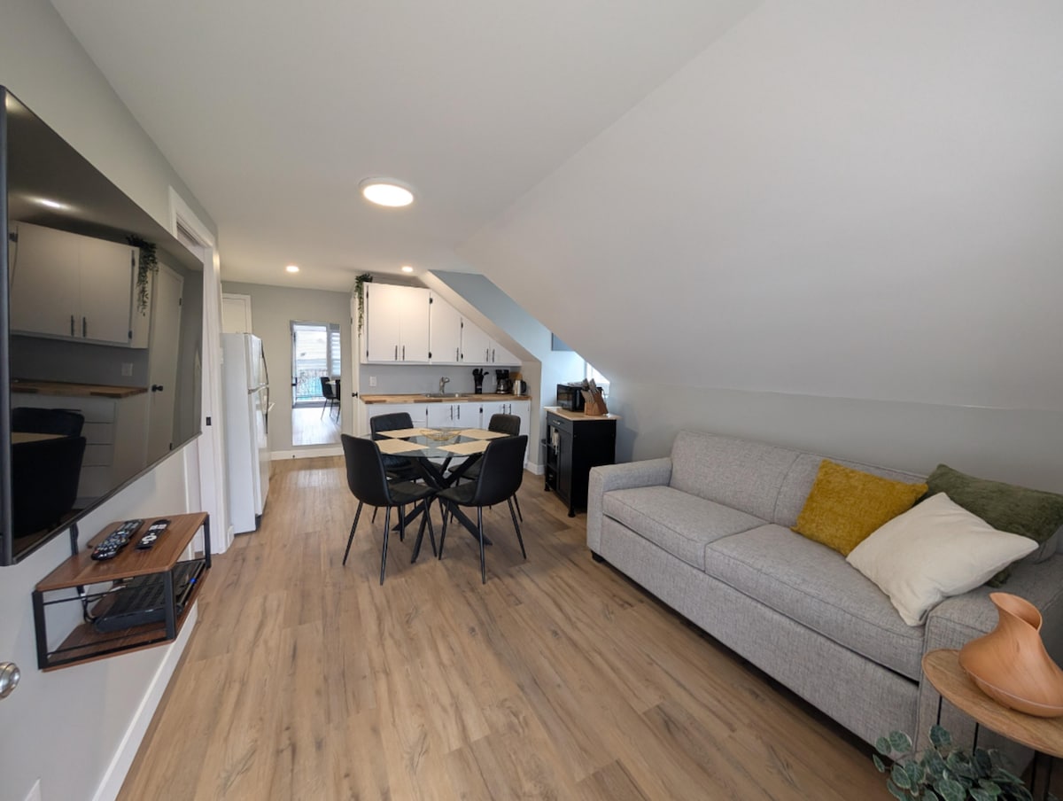 A light-filled living area features a comfortable gray sofa adorned with a yellow accent pillow. A round dining table with black chairs is positioned nearby, while modern kitchen appliances are visible in the background against a backdrop of soft, neutral tones.