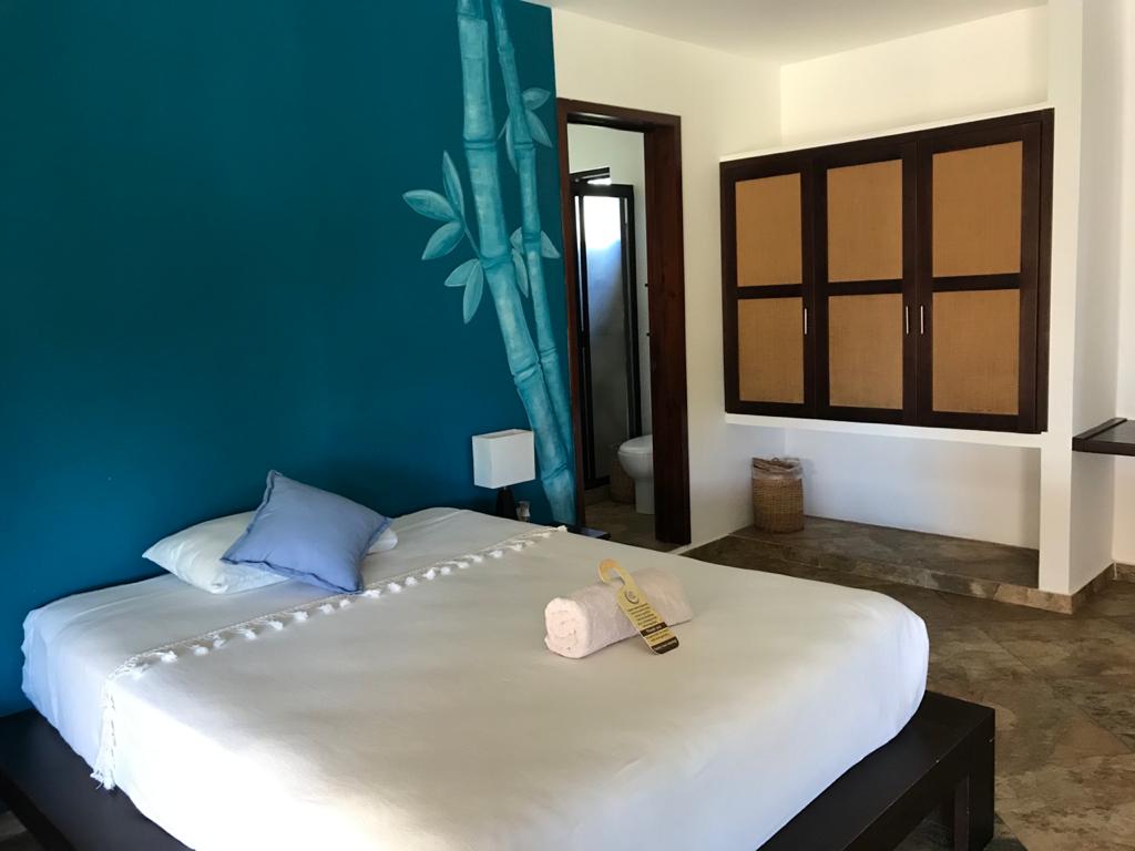 A spacious sleeping area features a white bed with a light blue pillow, complemented by a decorative accent wall in teal. A wooden cabinet with sliding doors is visible in the background, along with a small lamp and an organized bathroom area beyond.