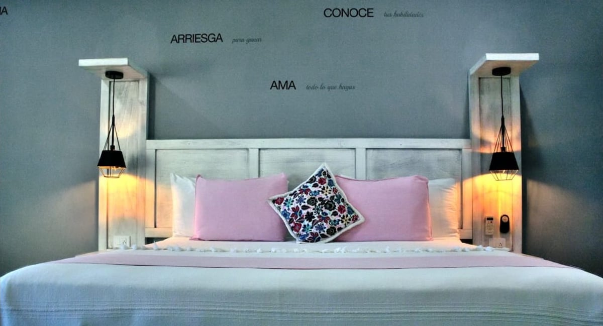 A spacious bed is centered against a grey wall, adorned with a decorative pillow featuring colorful floral patterns. Minimalist wooden nightstands with pendant lights offer soft illumination on either side, enhancing the contemporary coastal decor.