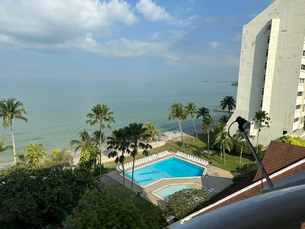 An aerial view captures a serene coastal landscape featuring a swimming pool bordered by lounge chairs. Lush palm trees surround the area, leading to a sandy beach that meets the calm sea, under a partly cloudy sky.