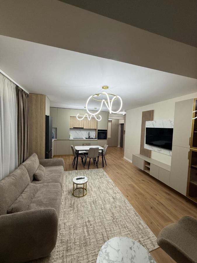 A stylish living area features a soft sofa and a modern coffee table, with light hardwood flooring. A contemporary light fixture hangs above, enhancing the space's ambiance. The kitchen is visible in the background, showcasing integrated appliances, while a TV is mounted on an attractive media unit.