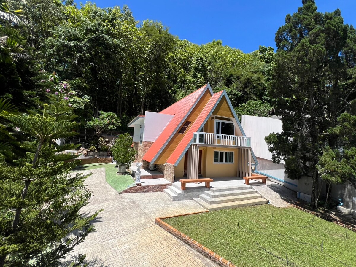 An A-frame cabin is set within a lush green landscape, surrounded by trees. The structure features a red roof and yellow walls, with a spacious outdoor area made of stone tiles and grassy patches, providing ample space for relaxation.