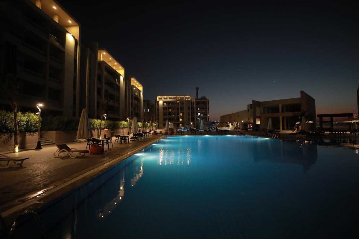 An expansive pool area is seen at night, illuminated by soft lighting from surrounding buildings. The clear blue water reflects the glow of nearby lights. Lounge chairs and umbrellas are arranged along the poolside, creating a serene environment for relaxation.