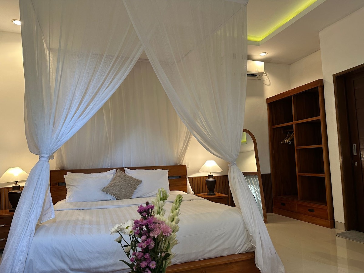 A serene bedroom is displayed with a spacious bed draped in sheer white curtains. Soft lighting is provided by bedside lamps. A small arrangement of fresh flowers is positioned on the floor, while a wooden wardrobe and a full-length mirror are visible in the background.