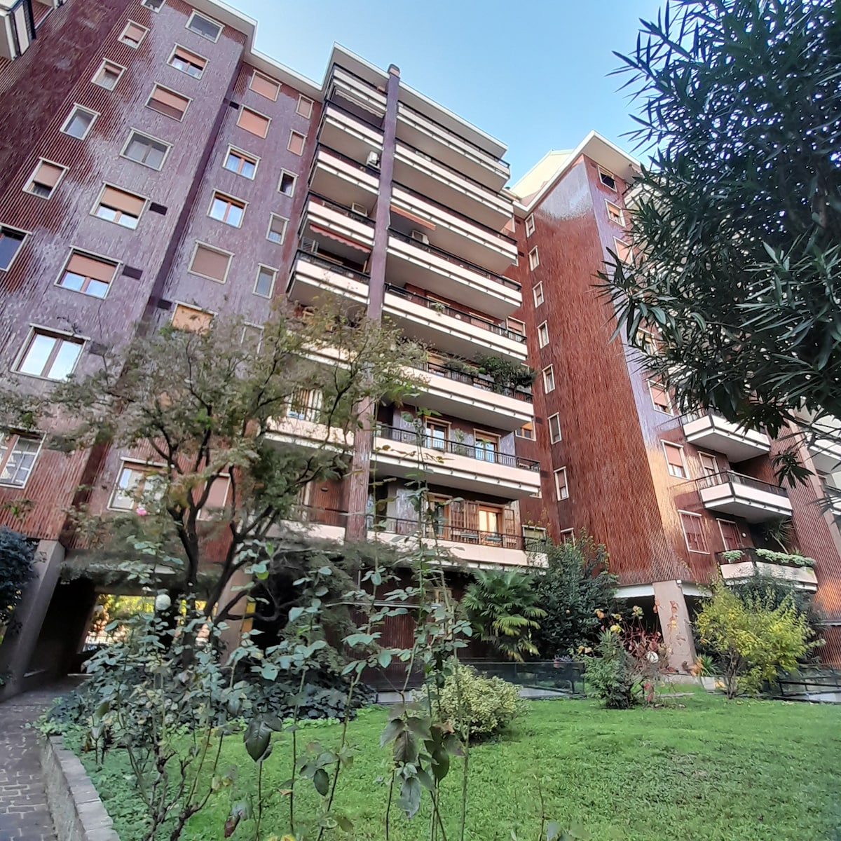 A multi-story building features a modern architectural design with wooden paneling. Balconies are visible, showcasing greenery and potted plants. The surrounding area includes a well-maintained garden with trees and shrubs, providing a tranquil environment.