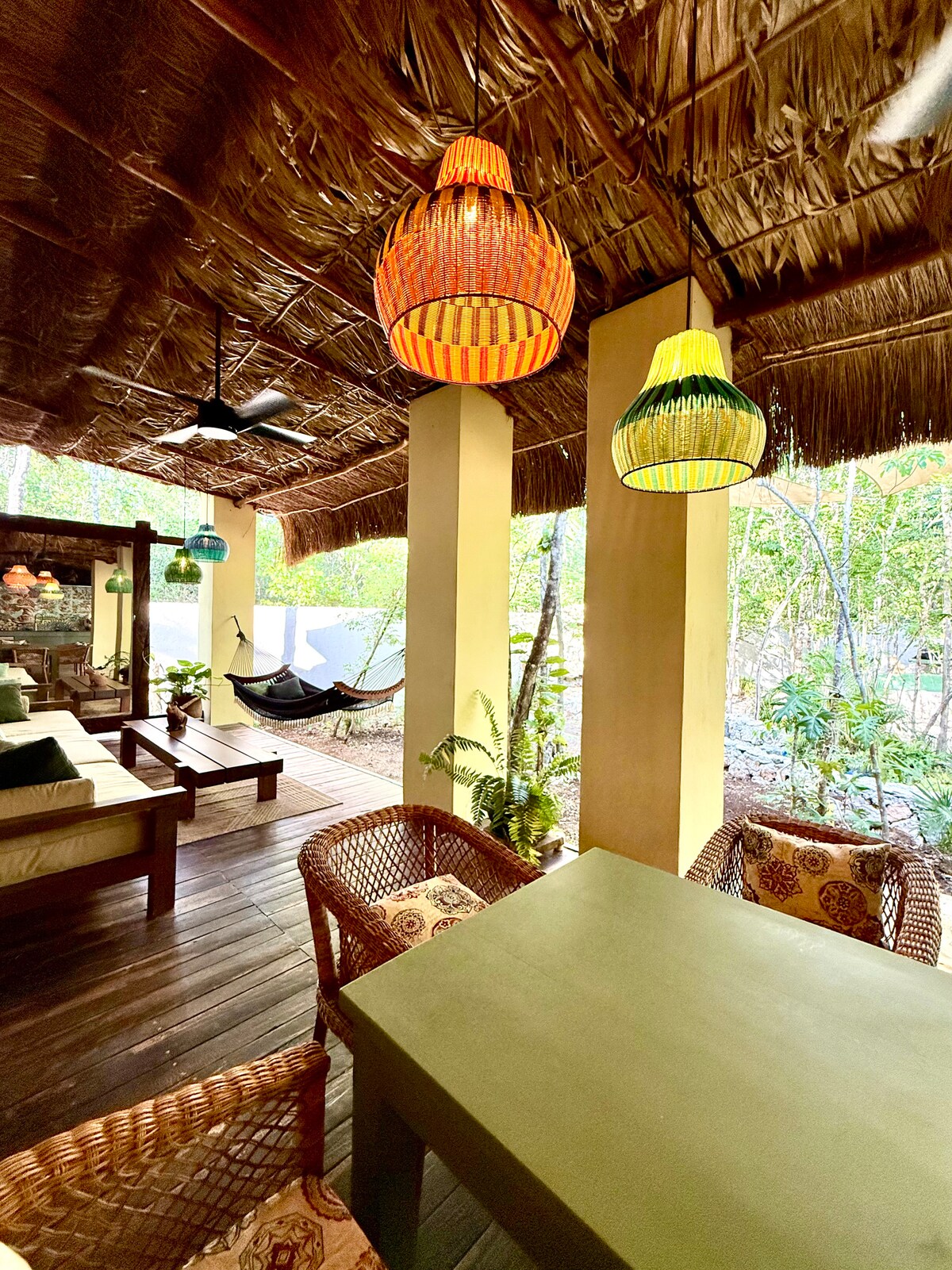 A covered outdoor seating area features wooden furniture and woven chairs, with a green table at the center. Colorful pendant lights hang from the thatched ceiling above, adding vibrancy. A hammock is visible in the corner, surrounded by lush greenery.