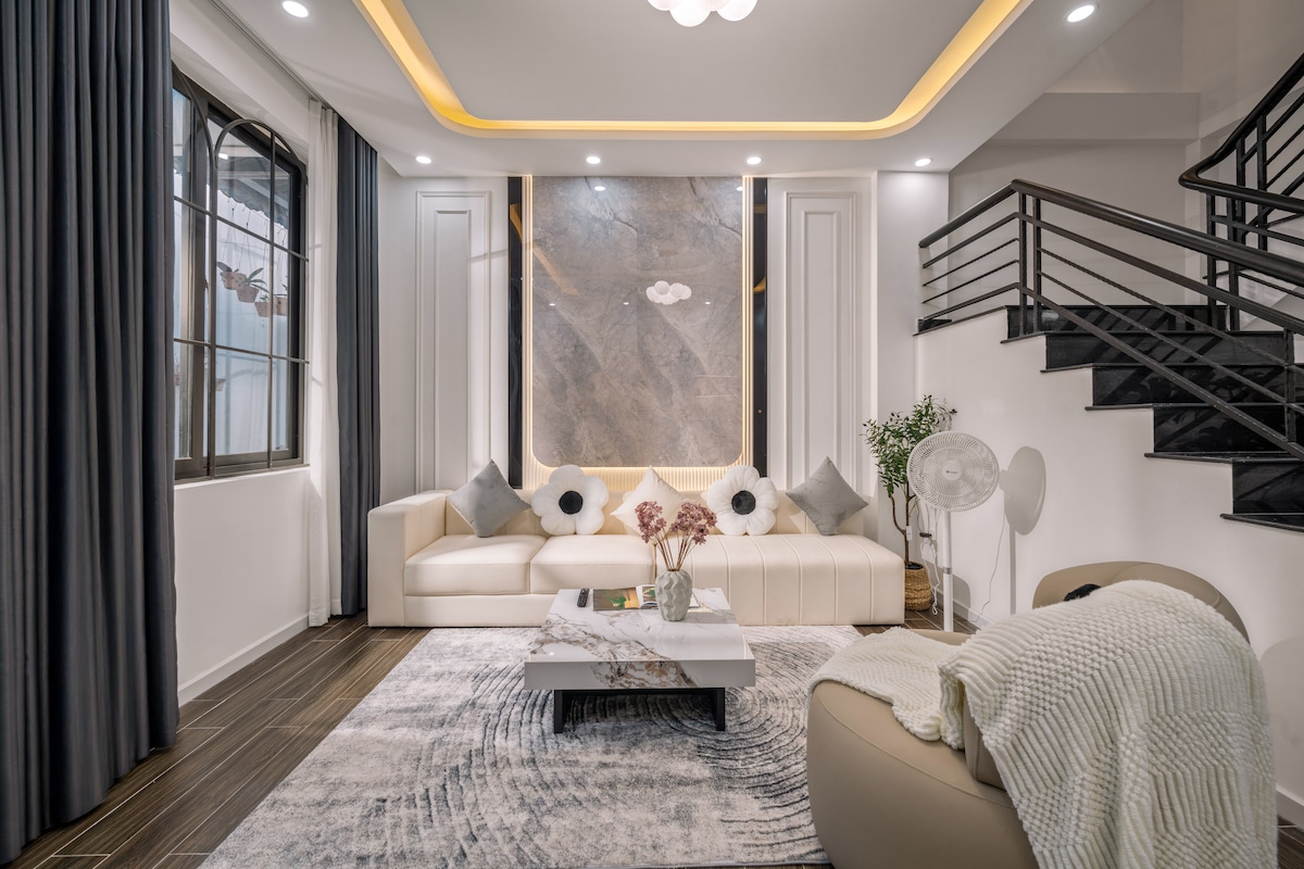 The living area includes a light-colored sectional sofa adorned with accent pillows. A central coffee table sits on a patterned area rug. Natural light enters through large windows, enhancing the space's brightness, while the staircase leads to the upper level.