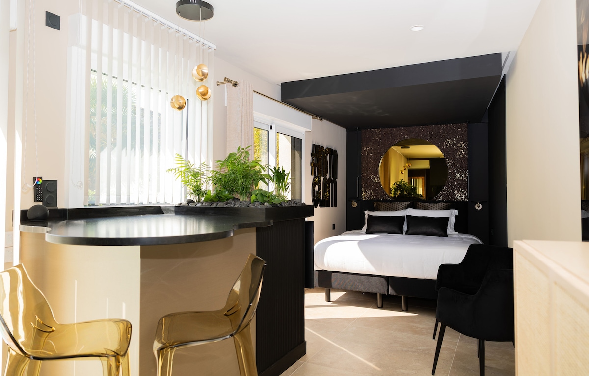 A modern studio features a spacious bed with elegant bedding, softened by natural light from large windows. A unique bar area with sleek chairs is complemented by indoor plants, providing a touch of greenery. The design reflects a blend of sophistication and functionality.