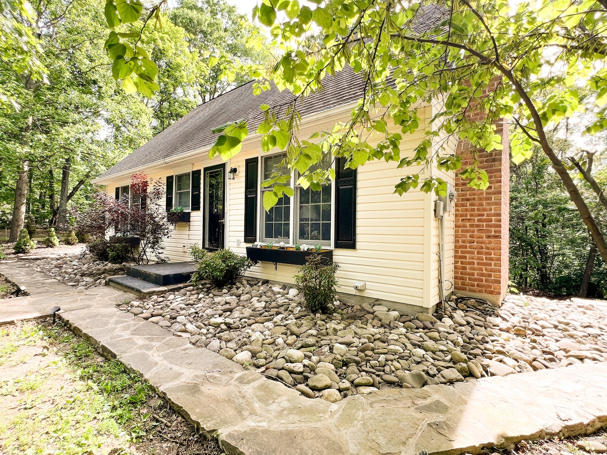 The exterior of the retreat is framed by greenery, featuring a light-colored facade with dark shutters. A stone walkway leads up to the entrance, accompanied by landscaped areas with smooth pebbles and a mix of plants. Natural light highlights the inviting atmosphere of the home.