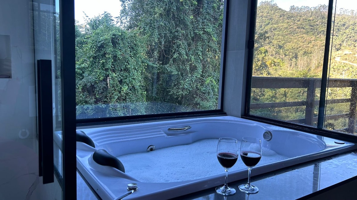 A large whirlpool bathtub is situated beside a window overlooking lush greenery. Two glasses of red wine are placed on the adjacent counter, complementing the serene view of the surrounding landscape. The space is bright and inviting, with natural light streaming through.
