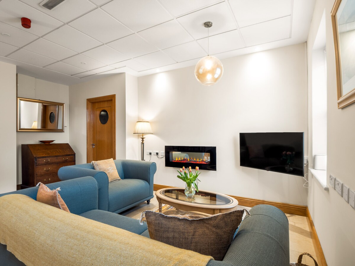 A spacious living area is seen, featuring two comfortable sofas in shades of blue and beige. An electric fire is mounted on the wall, providing warmth. A coffee table centers the room, complemented by fresh flowers. A mirror and a wooden dresser add character.