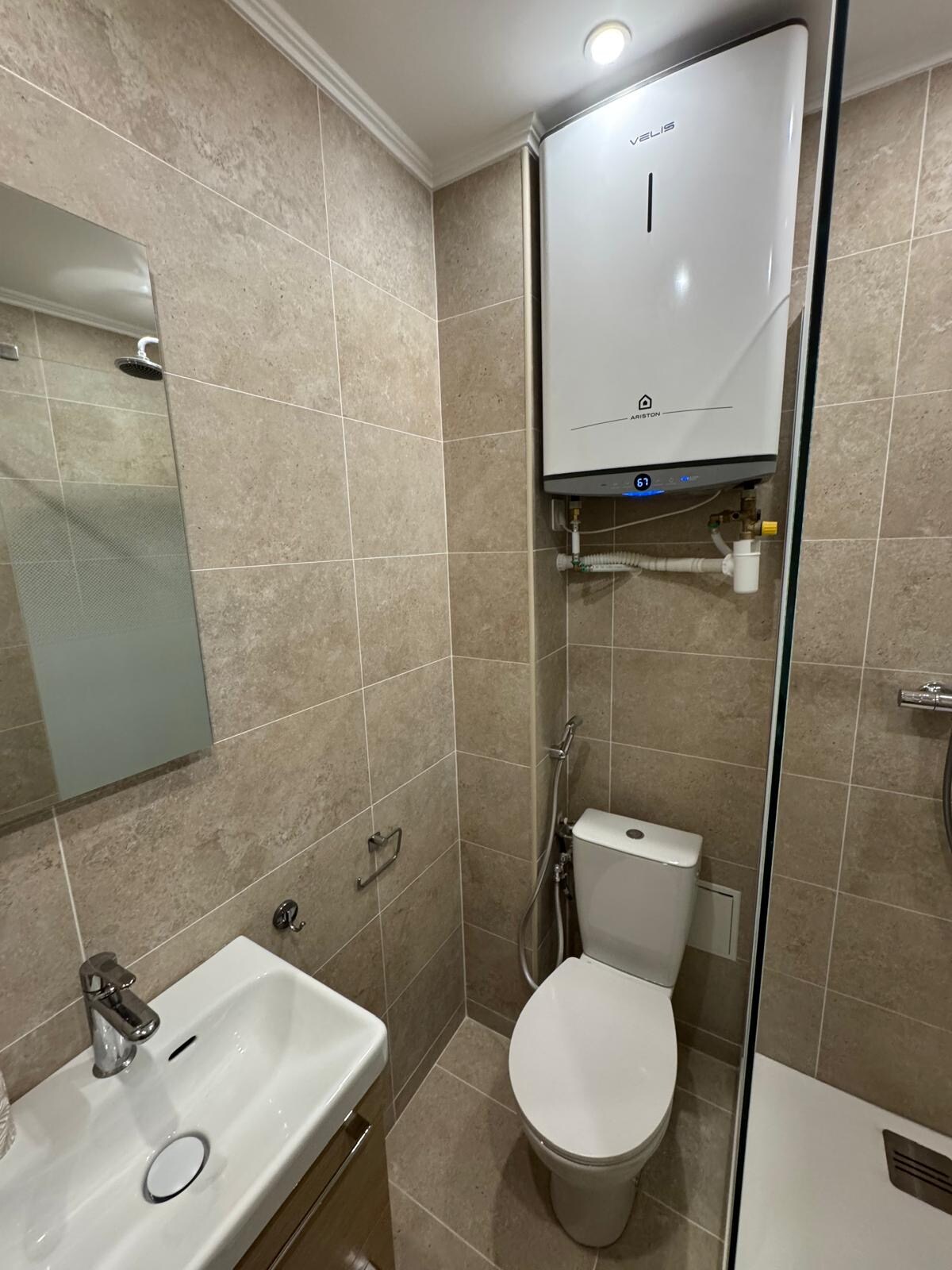 A modern bathroom features neutral-toned tiles covering the walls and floor. A mirror is mounted above a compact sink with a sleek faucet. A toilet is positioned beside a glass-enclosed shower area, with essential amenities visibly arranged for convenience.