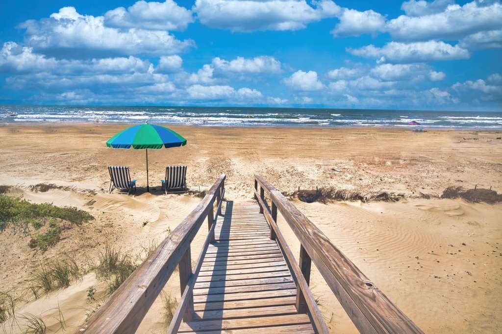 A wooden boardwalk leads to a sandy beach, with two beach chairs positioned under a colorful umbrella. The expansive ocean stretches out into the distance, where gentle waves meet a cloudy blue sky, creating a serene coastal atmosphere.