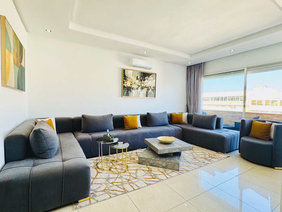 A spacious and modern living room features a large L-shaped sectional sofa adorned with cushions. A rectangular coffee table sits on a textured rug, complemented by light streaming through large windows. A piece of abstract wall art hangs above, adding a touch of elegance.